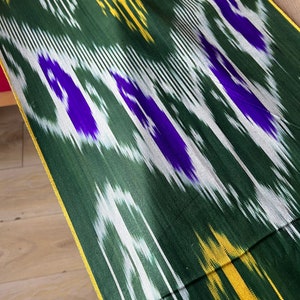 May include: Close-up of a green fabric with a vibrant ikat pattern. The design features white, yellow, and purple accents, creating a dynamic visual texture. The fabric has a gold border.