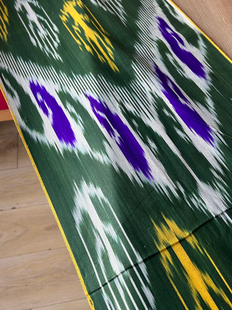 May include: Close-up of a green fabric with a vibrant ikat pattern. The design features white, yellow, and purple accents, creating a dynamic visual texture. The fabric has a gold border.