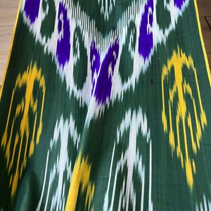 May include: Green fabric with a repeating pattern of white, yellow, and purple shapes. The fabric has a slight sheen and is folded, showing the texture and design. The edges are trimmed with a yellow border.