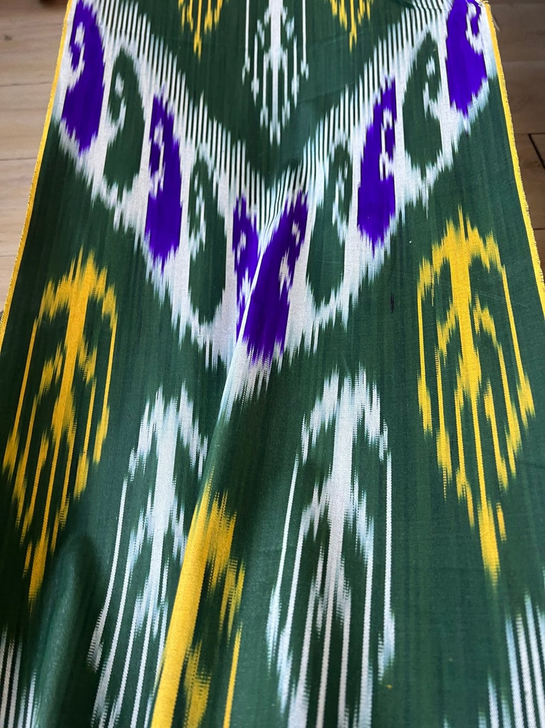 May include: Green fabric with a repeating pattern of white, yellow, and purple shapes. The fabric has a slight sheen and is folded, showing the texture and design. The edges are trimmed with a yellow border.