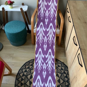 May include: A long purple and white ikat fabric with a yellow border. The fabric is draped over a chair and a small rug is on the floor.