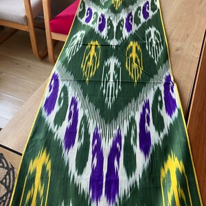 May include: A long, rectangular textile with a repeating pattern of white, purple, and yellow shapes on a dark green background. The edges are trimmed with a yellow border. The fabric is laid out on a wooden surface.