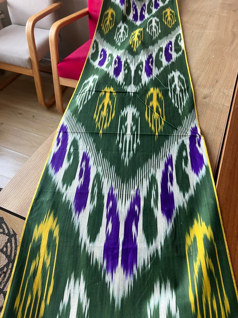 May include: A long, rectangular textile with a repeating pattern of white, purple, and yellow shapes on a dark green background. The edges are trimmed with a yellow border. The fabric is laid out on a wooden surface.