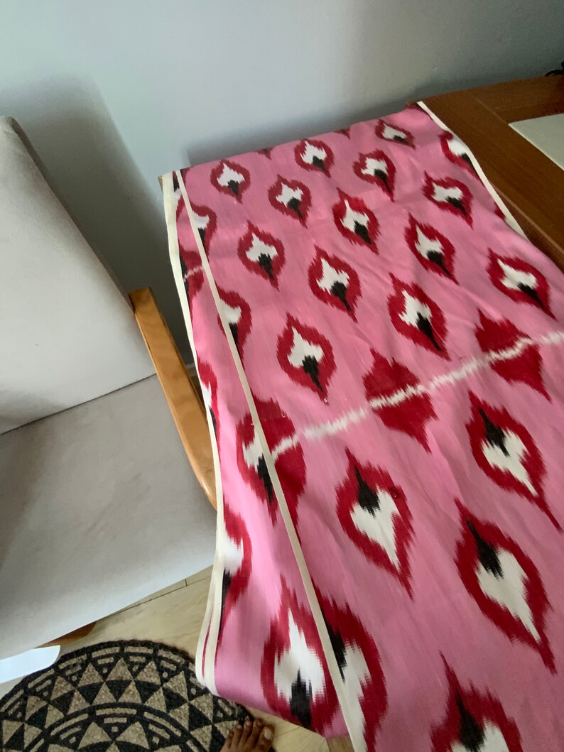 May include: Pink fabric with a white and black ikat pattern. The fabric has a white and black trim along the edges.