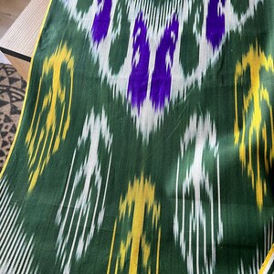 May include: Green fabric with a repeating pattern of white, yellow, and purple shapes. The fabric has a sheen and a yellow border. The pattern is a mix of vertical and diagonal lines and shapes.