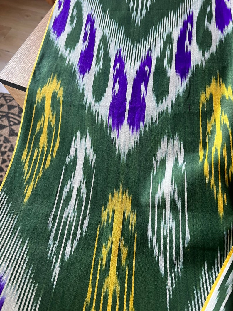 May include: Green fabric with a repeating pattern of white, yellow, and purple shapes. The fabric has a sheen and a yellow border. The pattern is a mix of vertical and diagonal lines and shapes.