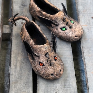 Peut inclure: Une paire de pantoufles en feutre marron en forme de dragons avec des ailes en feutre noir et des yeux verts et orange.