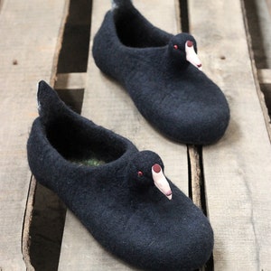 Op de afbeelding: Een paar zwarte wollen pantoffels in de vorm van eenden met rode ogen en roze snavels. De pantoffels liggen op een houten oppervlak.