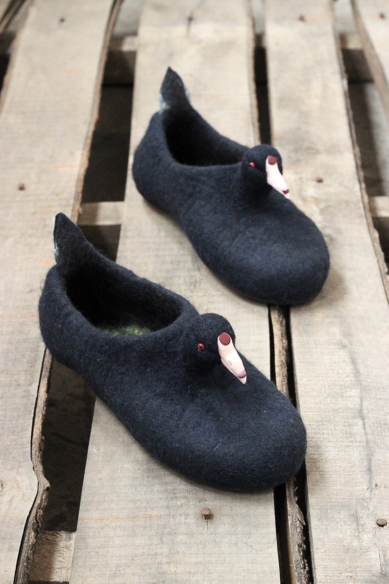 Op de afbeelding: Een paar zwarte wollen pantoffels in de vorm van eenden met rode ogen en roze snavels. De pantoffels liggen op een houten oppervlak.