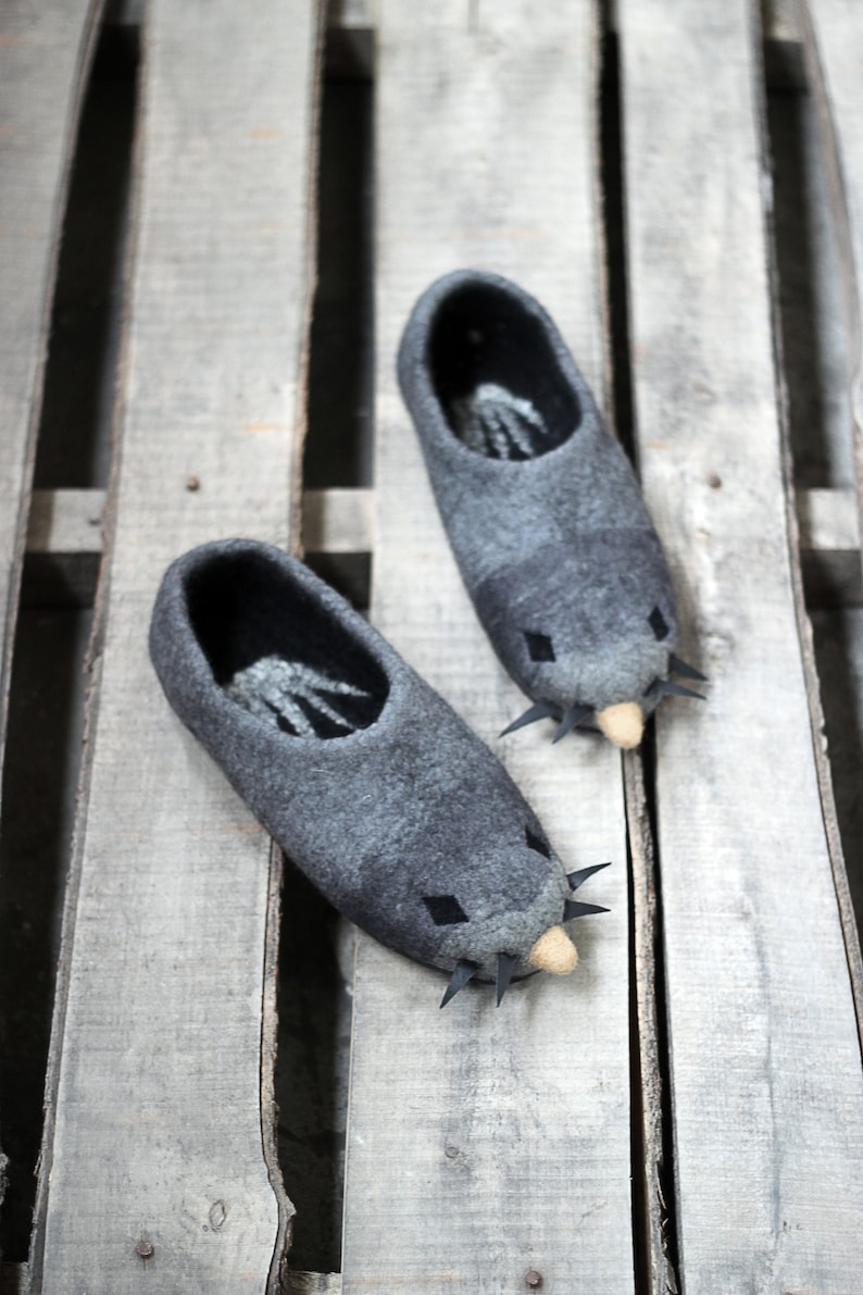 Peut inclure: Deux pantoufles en feutre gris en forme de taupe, avec des yeux en forme de losange noir et des d&eacute;tails pointus. Les pantoufles ont un nez beige clair et sont pos&eacute;es sur une palette en bois.