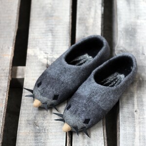 Peut inclure: Une paire de pantoufles grises en feutre en forme de souris, avec des moustaches noires et de petits nez beiges. Les pantoufles ont des yeux noirs en forme de losange et sont pos&eacute;es sur une surface en bois.