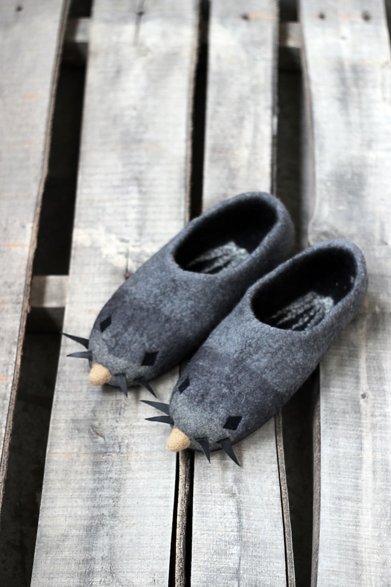 Peut inclure: Une paire de pantoufles grises en feutre en forme de souris, avec des moustaches noires et de petits nez beiges. Les pantoufles ont des yeux noirs en forme de losange et sont pos&eacute;es sur une surface en bois.