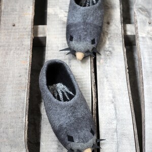 Peut inclure: Deux pantoufles en feutre gris en forme de taupe, avec des d&eacute;tails noirs pour les yeux et les moustaches, et un nez beige. Les pantoufles sont pos&eacute;es sur un fond de palette en bois. L'int&eacute;rieur des pantoufles pr&eacute;sente un motif noir et blanc.