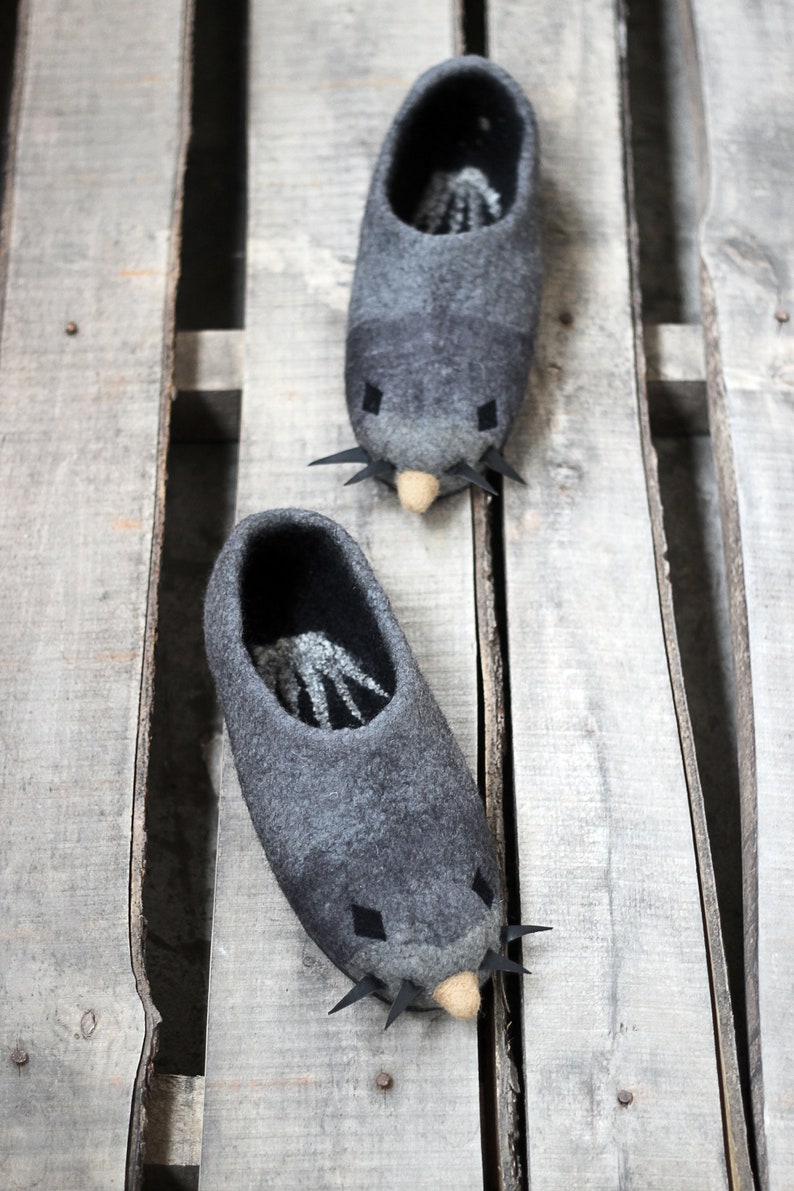 Peut inclure: Deux pantoufles en feutre gris en forme de taupe, avec des d&eacute;tails noirs pour les yeux et les moustaches, et un nez beige. Les pantoufles sont pos&eacute;es sur un fond de palette en bois. L'int&eacute;rieur des pantoufles pr&eacute;sente un motif noir et blanc.