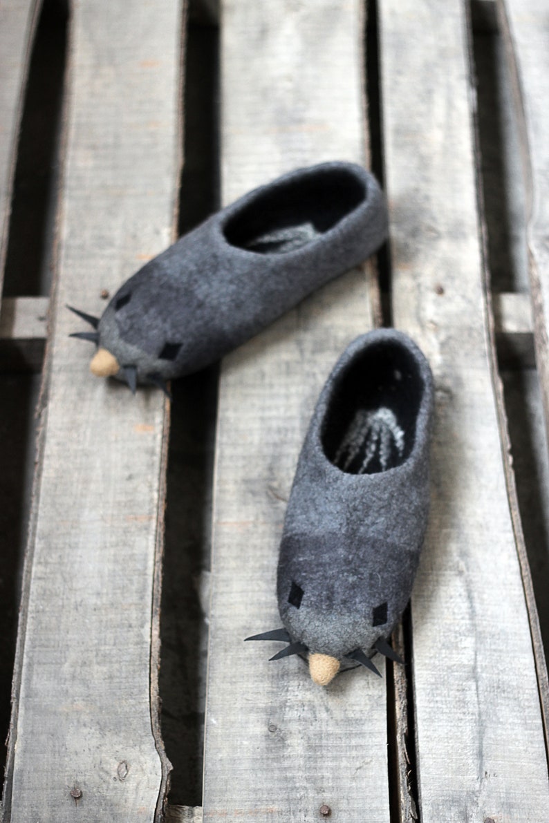Peut inclure: Une paire de pantoufles grises en feutre en forme de souris, avec des moustaches noires et de petits nez beiges. Les pantoufles sont pos&eacute;es sur une surface en bois vieilli, pour un design fantaisiste et confortable. Chaussons d'int&eacute;rieur.