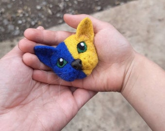 May include: A felt cat head with a blue and yellow design, representing the Ukrainian flag. The cat has green eyes and a black nose.