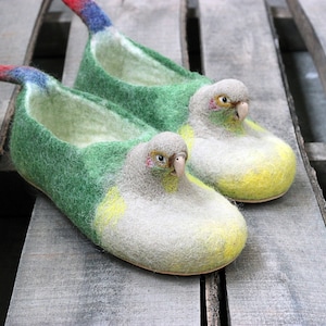 Op de afbeelding: Een paar groene en gele wollen pantoffels in de vorm van papegaaien. De pantoffels hebben een rood en blauw staartveer detail.