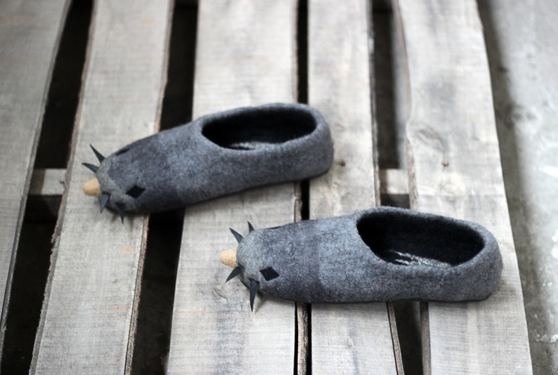 Peut inclure: Une paire de pantoufles en feutre gris en forme de taupe, avec des d&eacute;tails noirs en forme de losange et des pointes. Les pantoufles ont un int&eacute;rieur sombre et un nez brun clair. Pos&eacute;es sur une palette en bois.