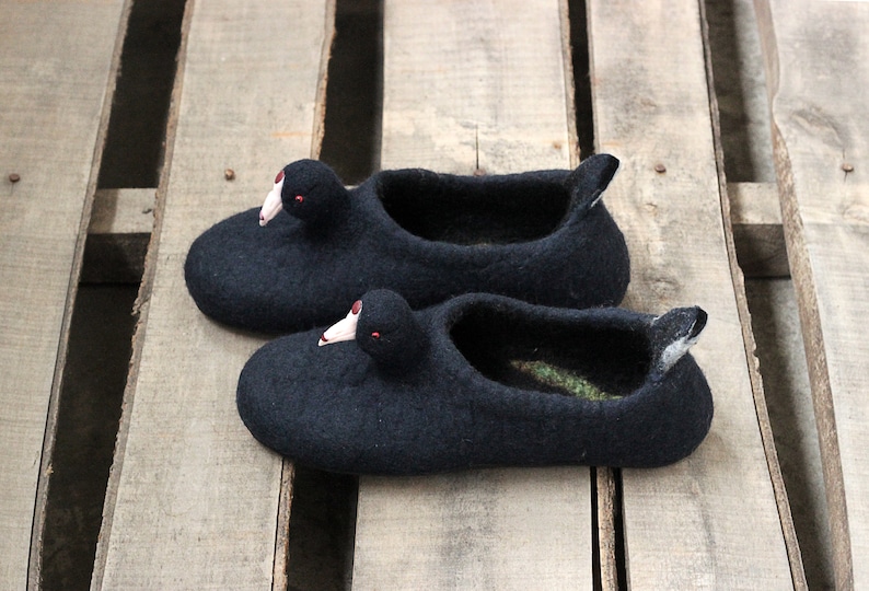 Op de afbeelding: Een paar zwarte wollen pantoffels in de vorm van een eend met rode ogen. De pantoffels liggen op een houten oppervlak.
