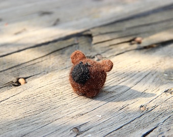 Pode incluir: Um pequeno urso de feltro marrom com um nariz e olhos de feltro preto. O urso está sentado em uma superfície de madeira.