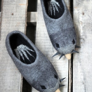 Peut inclure: Une paire de pantoufles en feutre gris avec un motif de taupe. Les pantoufles ont un int&eacute;rieur gris fonc&eacute; avec un motif blanc, des accents de diamants noirs et des pointes noires. Les pantoufles sont sur une surface en bois.