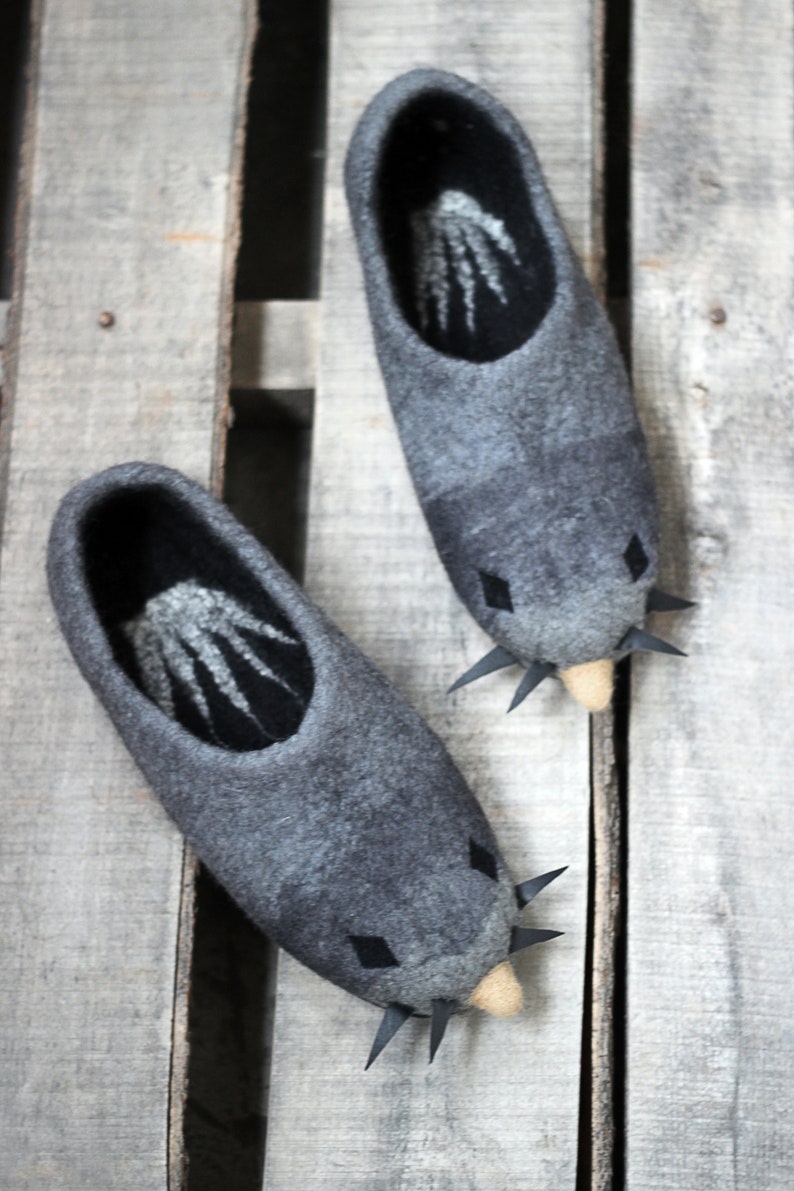 Peut inclure: Une paire de pantoufles en feutre gris avec un motif de taupe. Les pantoufles ont un int&eacute;rieur gris fonc&eacute; avec un motif blanc, des accents de diamants noirs et des pointes noires. Les pantoufles sont sur une surface en bois.
