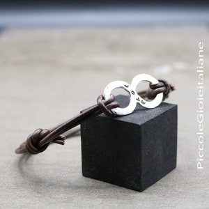 Pode incluir: Um bracelete de couro castanho com um amuleto de prata em forma de símbolo de infinito. O amuleto tem os números "10.4.17" gravados.
