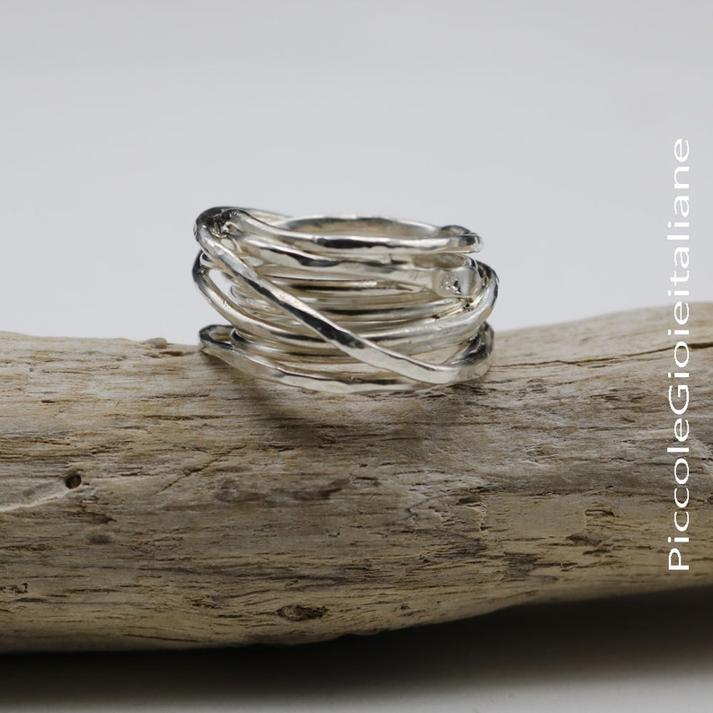 Op de afbeelding: Een zilveren ring met een uniek, gelaagd ontwerp. De ring heeft meerdere banden van verschillende breedtes die in elkaar grijpen om een ​​gestructureerd, opvallend stuk te cre&euml;ren. De ring wordt gepresenteerd op een stuk drijfhout.