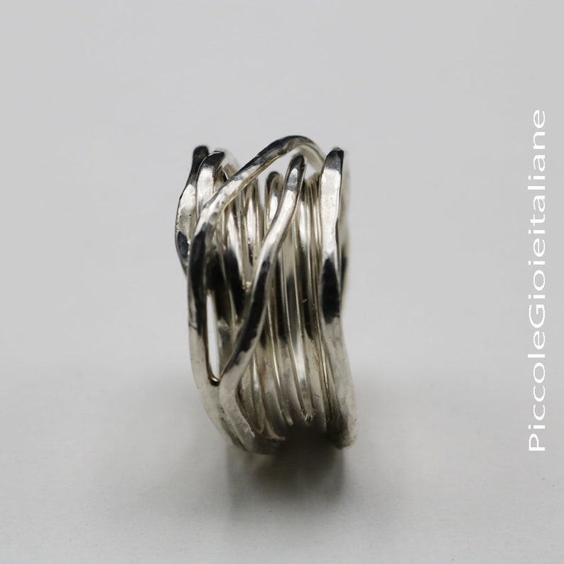 Op de afbeelding: Een zilveren ring met een uniek, geweven ontwerp. De ring heeft meerdere strengen metaal die in elkaar grijpen om een ​​gestructureerd, opvallend stuk te cre&euml;ren. Het ontwerp van de ring is modern en artistiek, geschikt voor dagelijks gebruik of speciale gelegenheden.