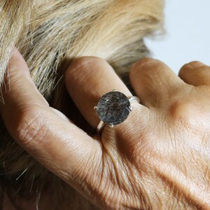 May include: A silver ring with a round, dark gray gemstone. The gemstone has a cloudy, textured appearance.