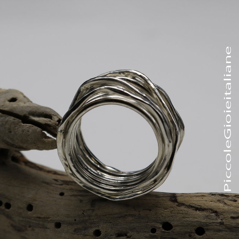 Op de afbeelding: Een zilveren ring met een textuur, golfachtig ontwerp. Het oppervlak van de ring heeft een gerimpeld uiterlijk, met meerdere banden die een gelaagd effect cre&euml;ren. De ring staat tegen een neutrale achtergrond.