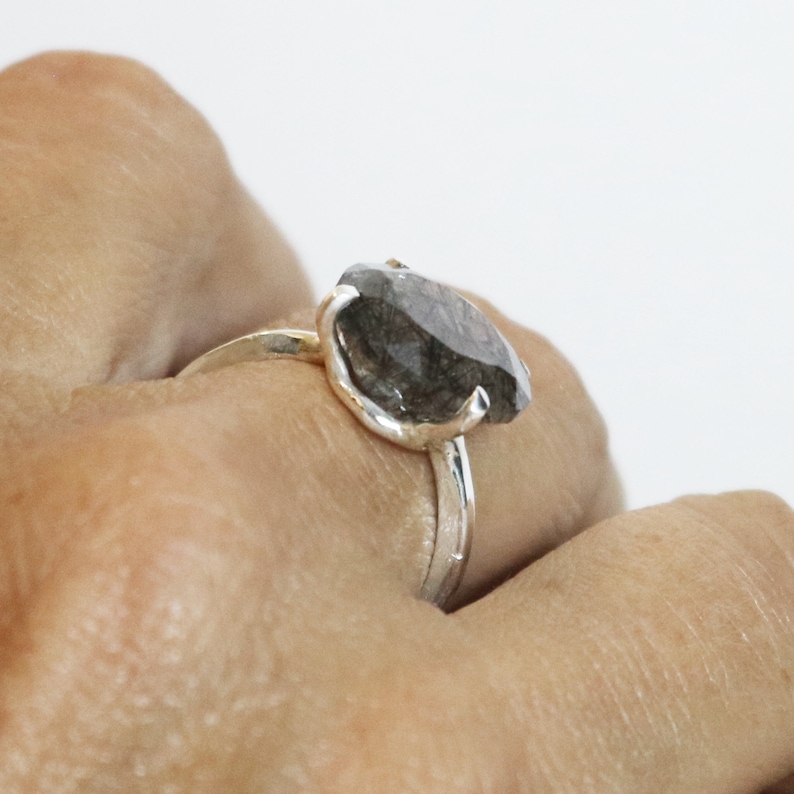 May include: A silver ring with a large, dark gray, faceted gemstone. The ring is worn on a person's finger.