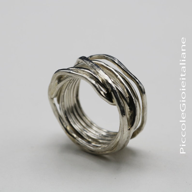 Op de afbeelding: Een zilveren ring met een uniek ontwerp. De ring heeft meerdere strengen metaal die in elkaar verweven zijn om een ​​gestructureerde, organische uitstraling te cre&euml;ren. De ring staat tegen een witte achtergrond.