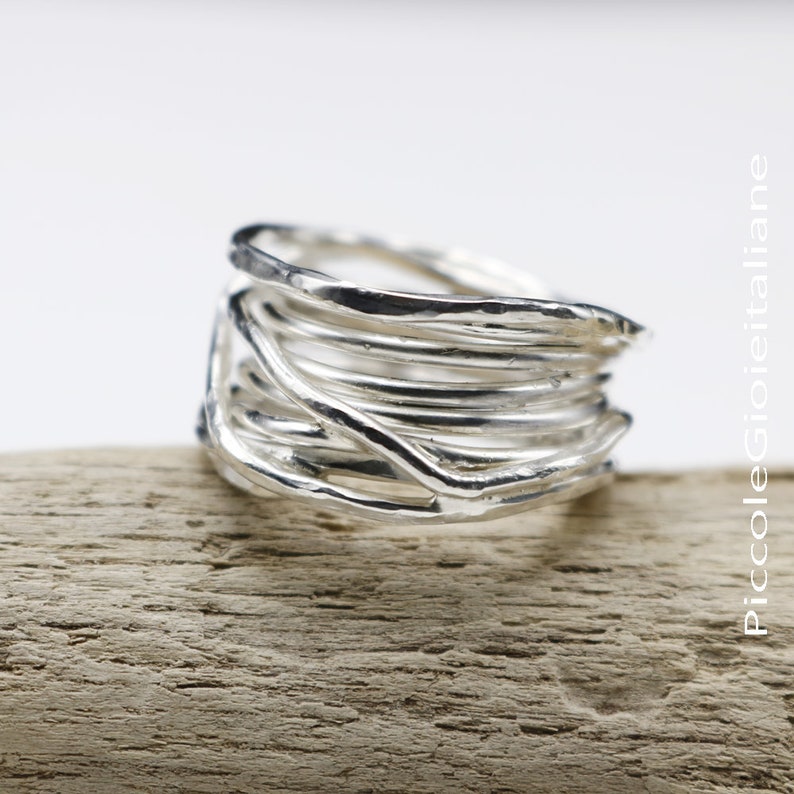 Op de afbeelding: Een zilveren ring met een uniek ontwerp. De ring heeft meerdere strengen zilverdraad die gewikkeld en verweven zijn, waardoor een textuurrijk, artistiek uiterlijk ontstaat. De ring wordt getoond op een stuk lichtbruin hout.