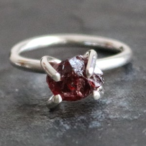 Può includere: Un anello d'argento con una pietra preziosa di granato rosso grezzo incastonata al centro. L'anello è in argento sterling e ha un design semplice e minimalista.