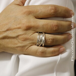 Op de afbeelding: Een zilveren ring met een geweven ontwerp. De ring heeft meerdere strengen zilver die met elkaar verweven zijn om een ​​gestructureerd, open uiterlijk te cre&euml;ren. De ring wordt aan een vinger gedragen en toont de ingewikkelde details en het vakmanschap.