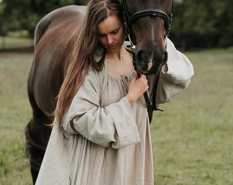 May include: A woman in a long, light brown linen dress is standing in a field with a brown horse. She is holding the horse's reins and looking down at the horse.