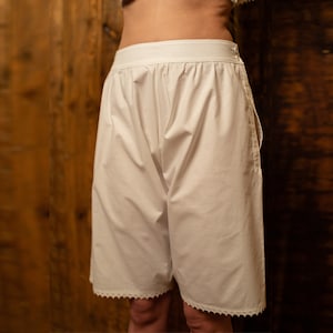 Könnte beinhalten: Weiße Baumwollshorts mit Spitzenbesatz. Die Shorts haben einen breiten Bund und eine lockere Passform.