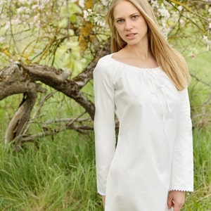 Pode incluir: Uma mulher com cabelo comprido e loiro vestindo um vestido branco de manga comprida com uma borda de renda. Ela est&aacute; em p&eacute; em um campo de grama verde com uma &aacute;rvore florida atr&aacute;s dela.