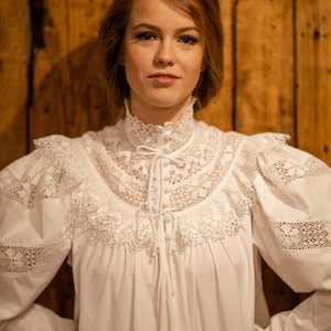 Puede incluir: Una blusa blanca de manga larga con un cuello alto de encaje y detalles de encaje en las mangas. La blusa tiene un lazo en el cuello y botones en la parte delantera.
