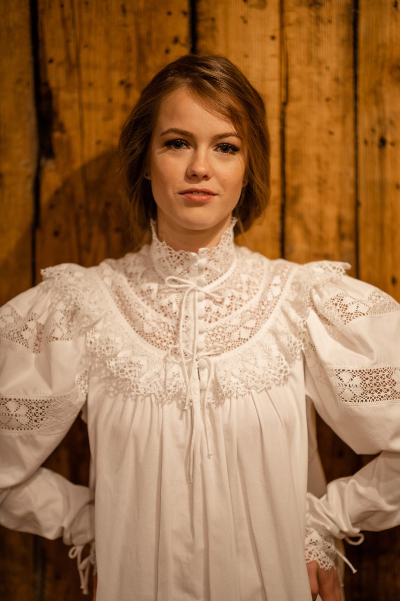 Puede incluir: Una blusa blanca de manga larga con un cuello alto de encaje y detalles de encaje en las mangas. La blusa tiene un lazo en el cuello y botones en la parte delantera.