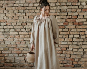 May include: A woman wearing a long, light brown linen dress with a gathered neckline and long sleeves. She is holding a woven basket and standing in front of a brick wall.