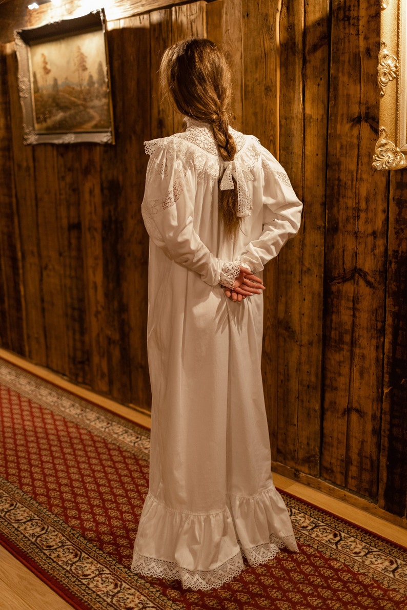 Puede incluir: Una mujer con un vestido blanco con ribete de encaje se encuentra en un pasillo con paneles de madera y una alfombra estampada. Su cabello est&aacute; trenzado y est&aacute; de espaldas a la c&aacute;mara.