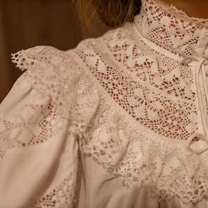 Puede incluir: Blusa blanca de encaje con cuello alto y cierre de botones. La blusa tiene un delicado patr&oacute;n de encaje y un cuello con volantes.