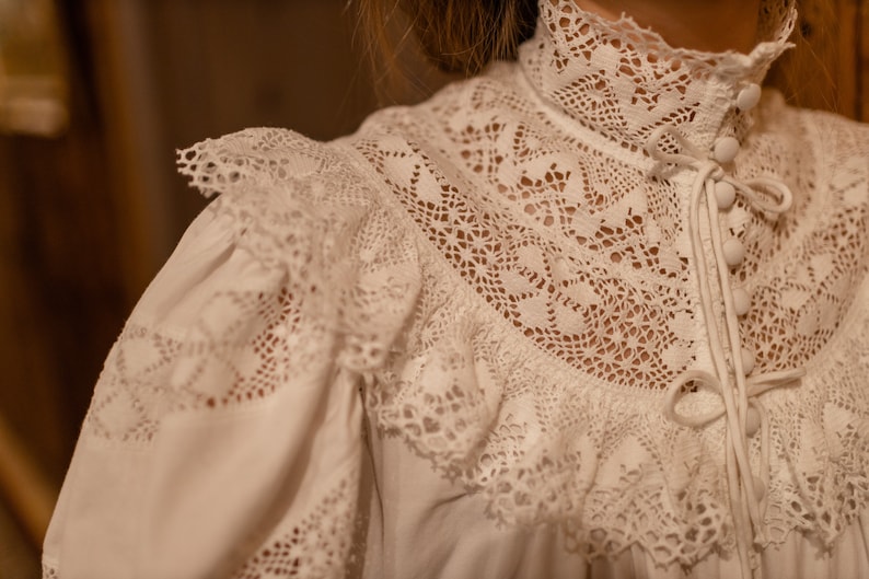 Puede incluir: Blusa blanca de encaje con cuello alto y cierre de botones. La blusa tiene un delicado patr&oacute;n de encaje y un cuello con volantes.