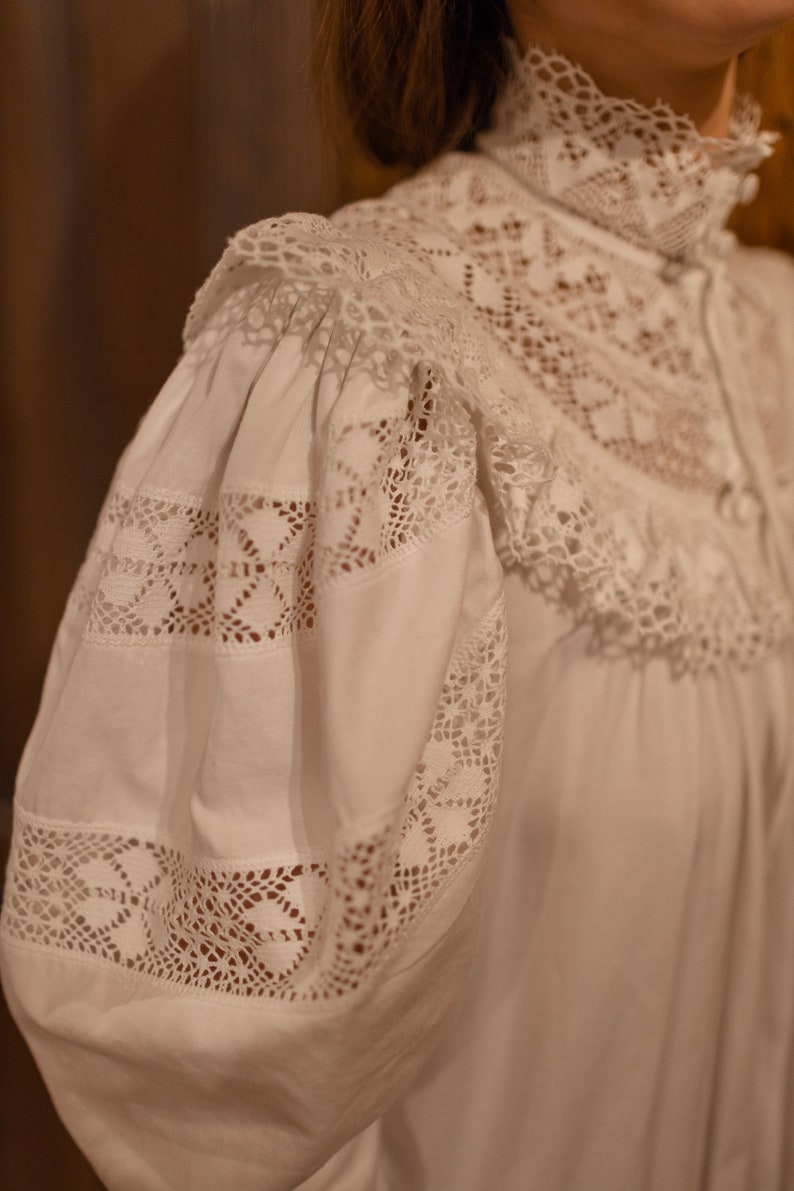 Puede incluir: Blusa blanca de algod&oacute;n con cuello alto de encaje y detalles de encaje en las mangas. El encaje es un patr&oacute;n delicado con un motivo floral.