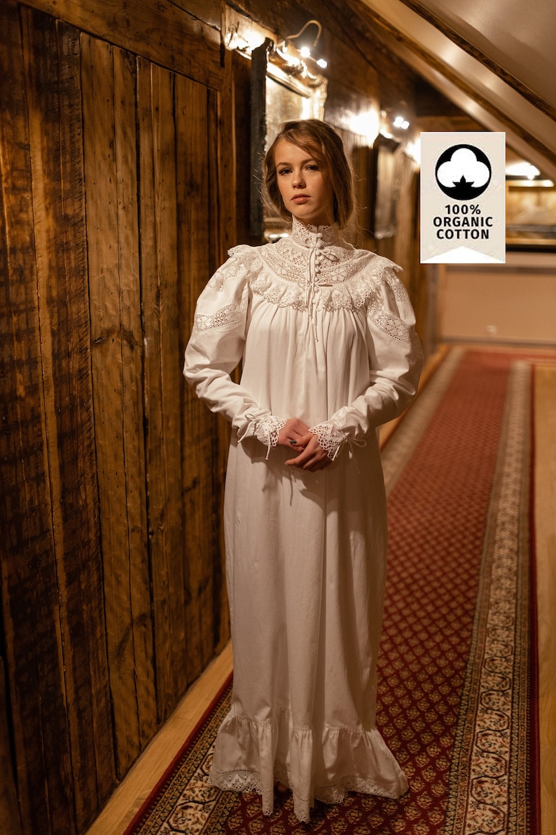 May include: A white nightdress with lace trim on the collar, sleeves, and hem. The nightdress features a high neck and a long, flowing design. The image includes a tag that reads "100% Organic Cotton".