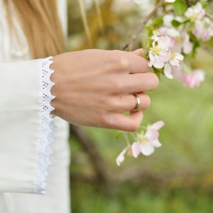 Pode incluir: Uma m&atilde;o de mulher com um anel de ouro estende-se para tocar um grupo de delicadas flores rosa e brancas num ramo.