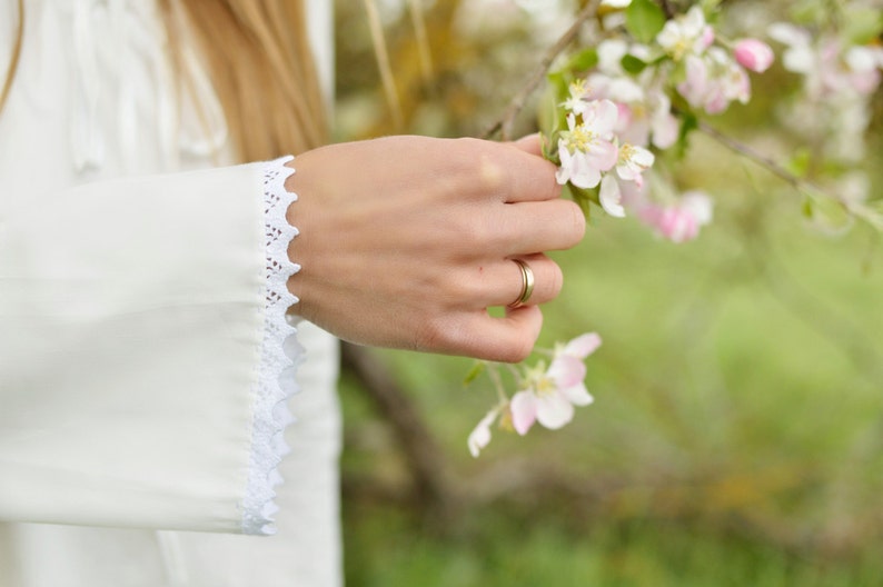 Pode incluir: Uma m&atilde;o de mulher com um anel de ouro estende-se para tocar um grupo de delicadas flores rosa e brancas num ramo.