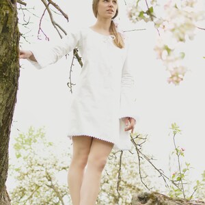 Pode incluir: Uma jovem com um vestido branco est&aacute; em um galho de &aacute;rvore em um pomar florido. Os galhos est&atilde;o cobertos de flores brancas.